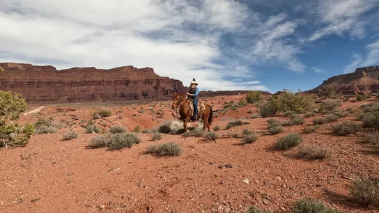 Person wearing a hat riding a brown horse across a red rocky desert landscape with sparse green shrubs and large red rock formations under a partly cloudy sky