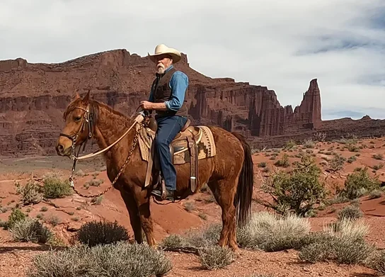 Man wearing a cowboy hat and vest sitting on a saddle riding a brown horse in a desert landscape with red rock formations and sparse vegetation