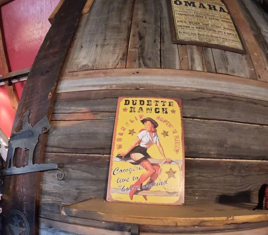 Yellow vintage-style metal sign depicting a pin-up cowgirl sitting on a fence with the text 'Dudette Ranch' and 'Cowgirls live to horse around' mounted on a rustic wooden wall