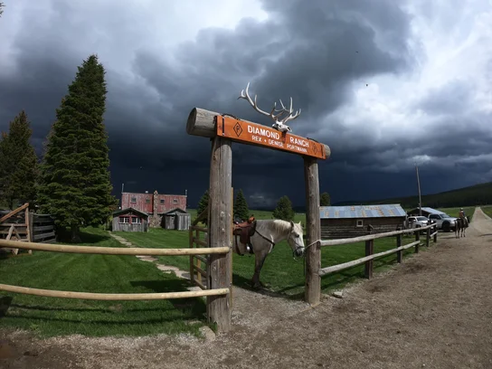 Saddled white horse tied to a wooden fence under an archway with an elk skull and antlers, with dark storm clouds overhead and rustic buildings in the background