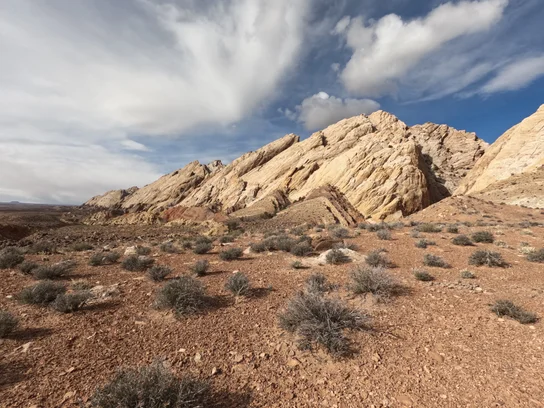 Desert landscape with reddish dirt and scattered low shrubs in the foreground, and layered, jagged tan rock formations under a partly cloudy blue sky