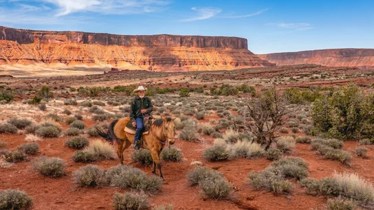 Man wearing a cowboy hat riding a horse through a desert landscape with red rock cliffs in the background