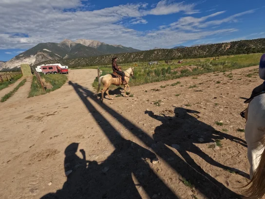 Person riding a light-colored horse on a dirt path with stacked hay bales and trailers in the background, surrounded by mountains and partly cloudy sky