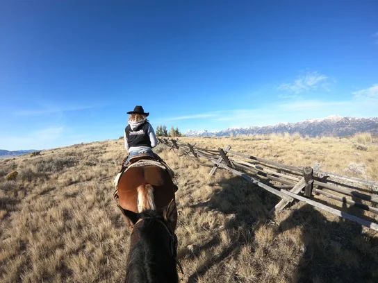 Person wearing a cowboy hat riding a horse along a grassy trail with a rustic wooden fence and snow-capped mountains in the distance under a clear blue sky