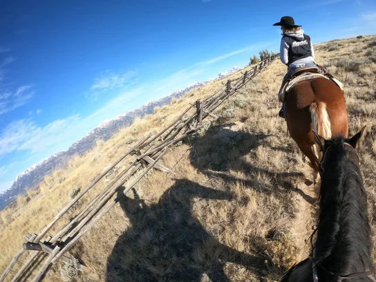 Person wearing a cowboy hat riding a brown horse along a dry grassy trail with a wooden fence on the side and snow-capped mountains in the distance under a blue sky