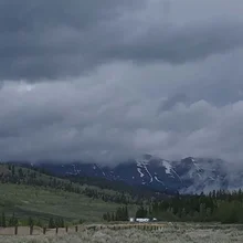 Cloud-covered mountain peaks with patches of snow behind green hills and trees under a gray overcast sky
