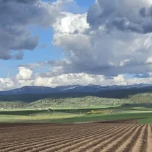 Plowed brown field with green hills, scattered buildings, and mountains under a cloudy sky