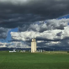Tall silo and several smaller storage tanks in a green field under a sky with dark storm clouds and patches of blue