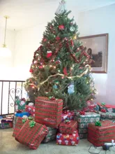 Fully decorated Christmas tree with lights, ornaments, a gold ribbon, and an angel tree topper surrounded by wrapped presents with festive bows