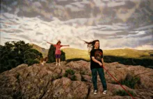 Two children standing on rocky terrain with one holding a red leash and a dog nearby under a cloudy sky