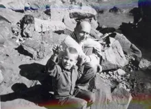 A child holding up a small object while sitting on rocks near a body of water with an adult crouching behind him
