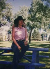 Person wearing a striped sleeveless shirt and a wide-brimmed black hat sitting on a blue picnic table in a park with trees in the background