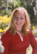 Smiling young woman with blonde hair wearing a red top and beaded necklace, outdoors with yellow flowers and greenery in the background