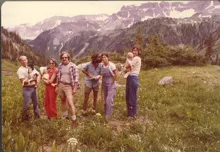 Group of six people standing in a meadow with wildflowers and mountains in the background, two holding babies and one holding a dog