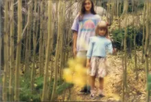 Two children standing on a narrow leaf-covered path surrounded by tall, thin trees with sparse branches
