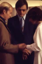 Three people standing closely together with heads bowed, holding hands in a moment of prayer or reflection
