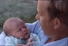 Adult holding a newborn baby wrapped in a light blue blanket with a concerned expression on the baby's face