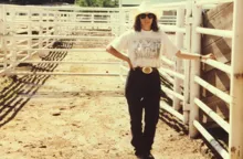 Person wearing a white hat, sunglasses, a white T-shirt with a print, black pants, and a large belt buckle standing on a dirt path between white metal rail fences