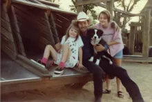 Two girls and a man wearing a cowboy hat smiling together on a wooden playground structure with a black and white dog