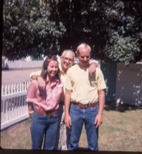 Three people standing closely outdoors with a white picket fence and leafy trees in the background, two younger adults flanking an older man who has his arms around them