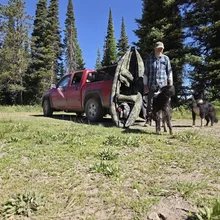 Man standing outdoors near a red pickup truck with two black and white dogs and a folded camouflage kayak hanging on the side of the truck