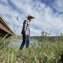 Man wearing a plaid shirt, jeans, and a cowboy hat standing near a river with green grass and plants in the foreground under a partly cloudy sky