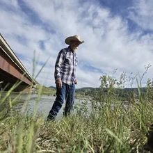 man wearing a cowboy hat and plaid shirt standing near a river with tall grass and a bridge on a partly cloudy day