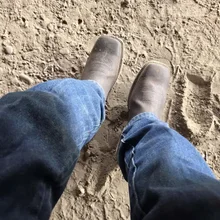 Person wearing worn brown cowboy boots and blue jeans standing on dirt ground with footprints and small rocks