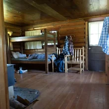 Rustic wooden cabin interior with bunk beds, wooden chairs, and clothes hanging on the chair backs and door