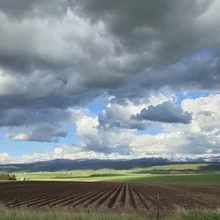 plowed farmland with rows leading toward a distant mountain range under a sky filled with dramatic dark and white clouds