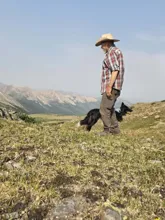 man in a cowboy hat and plaid shirt standing on a grassy hillside with a black and white dog among distant rocky mountains