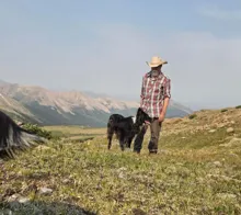 Man in a plaid shirt and cowboy hat standing on a grassy mountain slope with a black and white dog