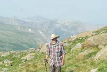 older man in a plaid shirt and wide-brimmed hat hiking on a rocky mountain slope