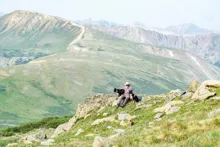 Person sitting on a rocky hillside with two dogs, surrounded by expansive green mountains under a clear sky