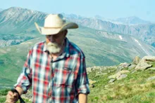 man in a plaid shirt and cowboy hat using a walking stick in a mountainous landscape