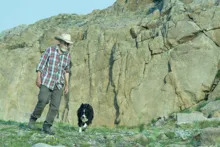 man in a plaid shirt and cowboy hat walking with a black and white dog near rocky terrain