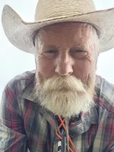 Elderly man with a full white beard wearing a straw cowboy hat and a plaid shirt, looking closely at the camera