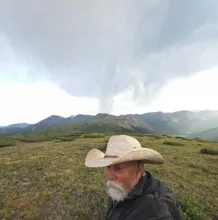 An older man with a white beard and a cowboy hat standing on a grassy hill with mountains and dark cloudy sky in the background