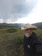 older man with a white beard wearing a wide-brimmed hat and dark jacket standing in a grassy mountainous landscape under a cloudy sky