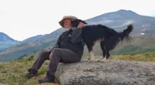 Man wearing a hat sitting on a large rock petting a black and white dog with mountains in the background