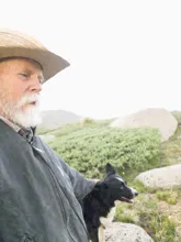 Older man with a white beard wearing a wide-brimmed hat and jacket sitting outdoors with a black and white dog in a rocky and green landscape