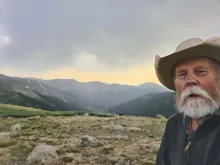 Older man with white beard and cowboy hat in a mountainous landscape under a cloudy sky at sunset