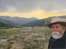 Older man with white beard and cowboy hat outdoors with distant mountains and a cloudy sky at sunset