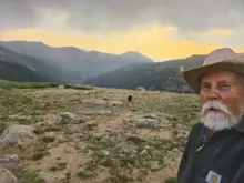 older man with white beard wearing a cowboy hat standing on a rocky mountain landscape at sunset