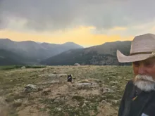 Older man with white beard and cowboy hat standing on rocky, grassy terrain with a dog in the distance and mountain range under a cloudy sky at sunset