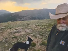 Bearded man wearing a large brimmed hat and dark jacket standing outdoors with a black and white dog on rocky terrain with mountains in the background during sunset