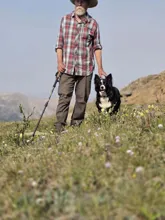 Man in a plaid shirt and hat standing on a grassy hillside with a black and white dog beside him