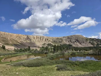Two small ponds surrounded by greenery with rocky hills and a partly cloudy blue sky in the background