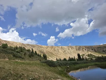 A mountain ridge with sparse trees and bushes, a grassy hillside, and a small body of water reflecting the cloudy sky