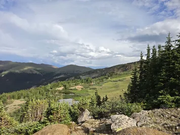 Rocky hillside with pine trees, a small pond, and distant rolling hills under a partly cloudy sky